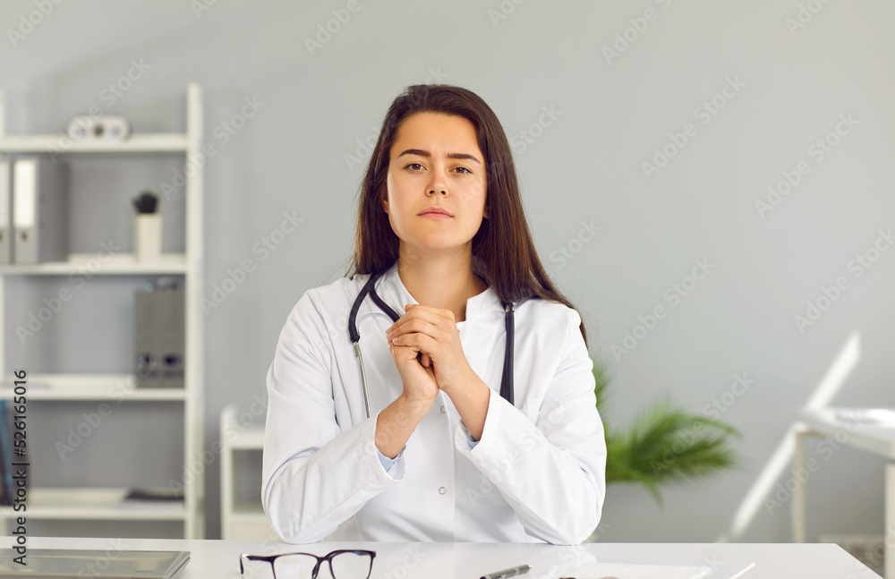 Portrait of online doctor during consultation via video call. Female physician listening to patient. Beautiful young woman in medical coat sitting at desk and looking at camera. Telemedicine concept Portrait of online doctor during consultation via video call. Female physician listening to patient. Beautiful young woman in medical coat sitting at desk and looking at camera. Telemedicine concept