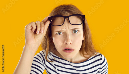 Confused woman looks at you with suspicious, doubtful and incredulous expression on her face. Close up of skeptical and suspicious young woman looking out from under glasses on orange background. photo