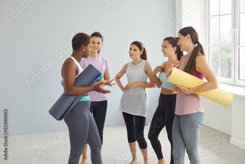 Multiracial group of happy fit beautiful young women having conversation after sports or yoga workout. Cheerful multiethnic international female friends with gym mats standing together and talking