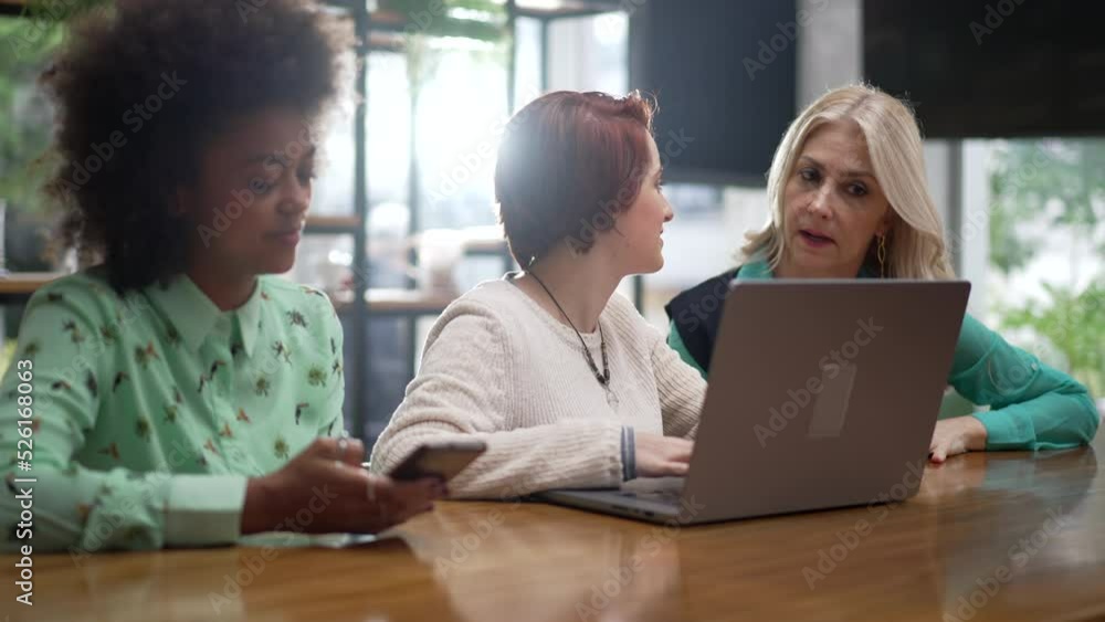 Three diverse women in front of laptop seated at coffee shop. Young employees with business woman executive coaching employees Three diverse women in front of laptop seated at coffee shop. Young employees with business woman executive coaching employees