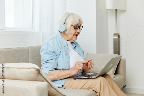 a happy modern old lady looks at the monitor holding a laptop on her lap talking through headphones smiling happily and gesticulating with her hands © Tatiana