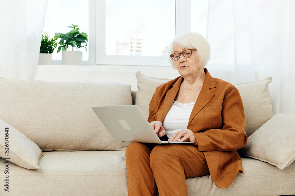 an attentive elderly woman with glasses is mastering new technologies using a laptop while sitting on the couch at home in a comfortable environment in a brown suit an attentive elderly woman with glasses is mastering new technologies using a laptop while sitting on the couch at home in a comfortable environment in a brown suit