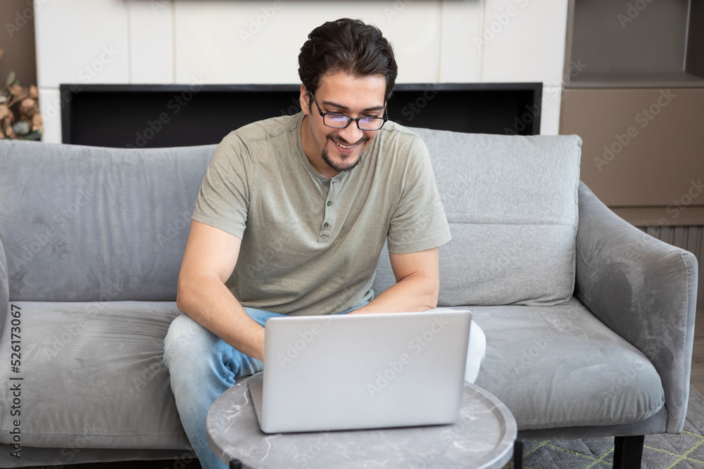Smiling handsome man sitting on the couch and working on laptop at home. Online education and distant work concept Smiling handsome man sitting on the couch and working on laptop at home. Online education and distant work concept