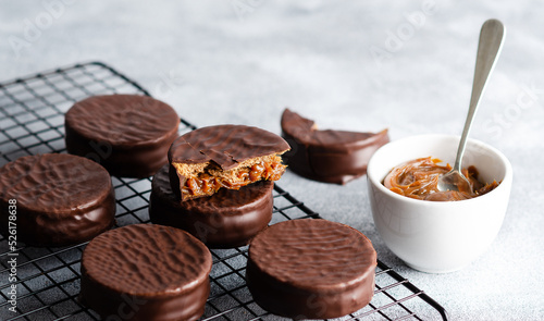 Dark chocolate alfajores on a dark rack and a bowl with dulce de leche, on grey background. 