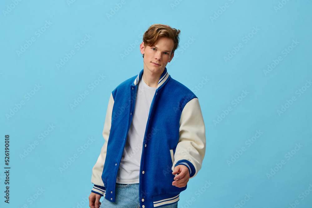 a handsome, attractive young man student in a trendy bomber jacket and jeans is standing friendly extending his hand to the camera on a light blue background. Horizontal photo with empty space