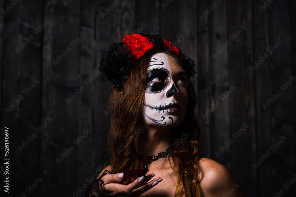 Portrait of a woman with makeup on her face in the form of a skull and with a wreath of red and black roses holding a dried rosebud in her hand. Portrait of a woman with makeup on her face in the form of a skull and with a wreath of red and black roses holding a dried rosebud in her hand.