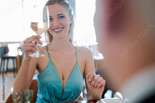 Eine junge, blonde Frau sitzt in ihrem hellblauen Abendkleid an einem festlich eingedeckten Tisch und unterhält sich freundlich mit ihrem Begleiter. Sie prostet ihm freundlich lächelnd zu. 