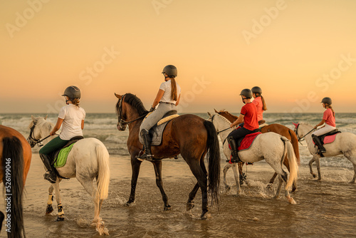 rear view of a group of 6 horses mounted by young riders trotting in the sea.