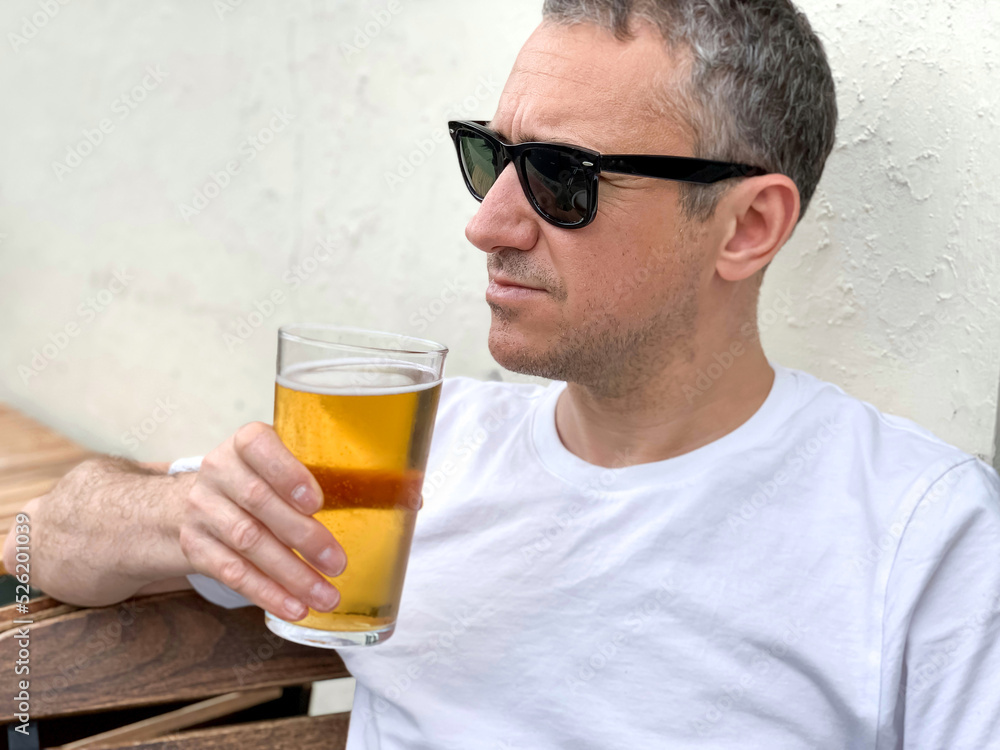 Middle aged man is drinking beer in a cafe or restaurant pub outdoor. Drinking alcohol. Candid. Refreshment, summer time, pub garden. wearing white t-shirt and sunglasses. Drinking lager or ale beer. Middle aged man is drinking beer in a cafe or restaurant pub outdoor. Drinking alcohol. Candid. Refreshment, summer time, pub garden. wearing white t-shirt and sunglasses. Drinking lager or ale beer.