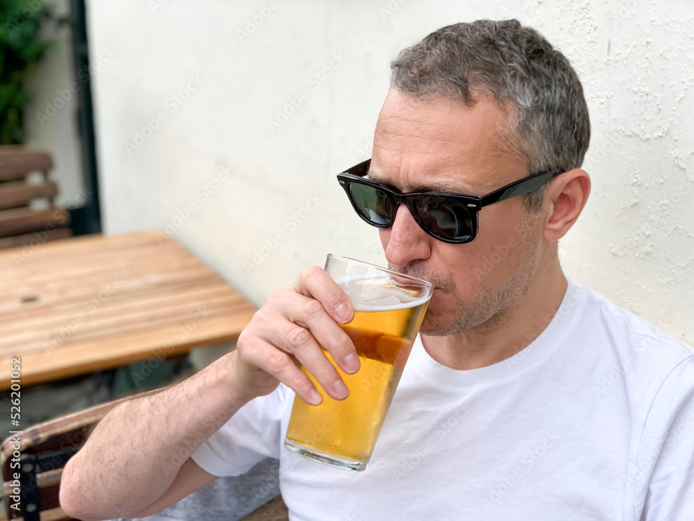 Middle aged man is drinking beer in a cafe or restaurant pub outdoor. Drinking alcohol. Candid. Refreshment, summer time, pub garden. wearing white t-shirt and sunglasses. Drinking lager or ale beer. Middle aged man is drinking beer in a cafe or restaurant pub outdoor. Drinking alcohol. Candid. Refreshment, summer time, pub garden. wearing white t-shirt and sunglasses. Drinking lager or ale beer.