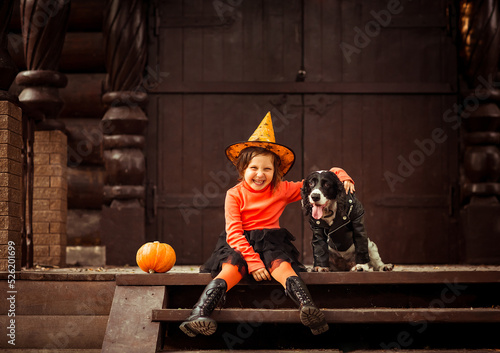 a small, beautiful girl in an orange and black witch costume and a hat, sitting on the porch with a dog in a leather jacket