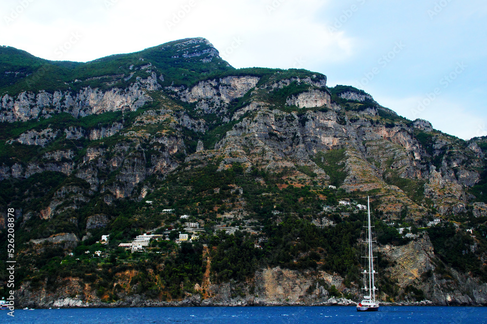 Daunting view in Amalfi at a huge mountain with rocky parts, greenery and scattered buildings while a white boat sits in the waters of the Tyrrhenian sea in the foreground under a cloudy azure sky