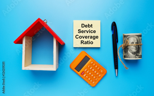 A picture house miniature, with fake money, Debt Service Coverage Ratio on notepad, calculator, and pen on blue background.