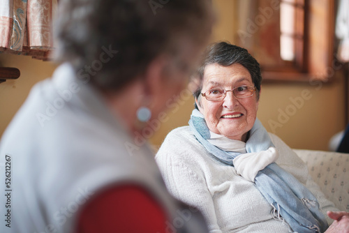 Happy elderly woman talking to friend sitting on sofa in retirement home having conversation