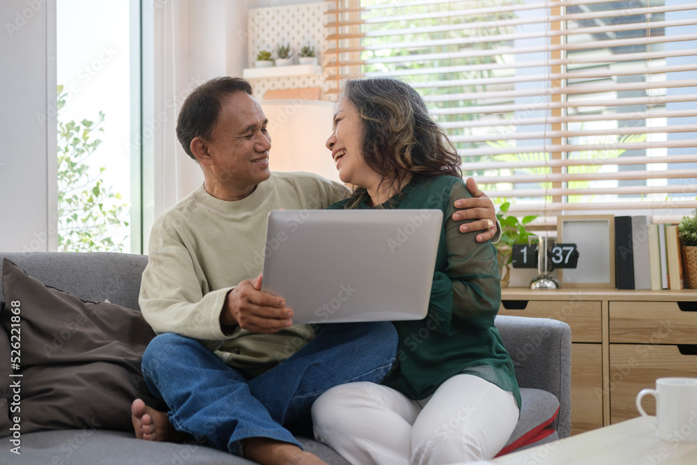 Happy senior couple sitting on couch and using laptop, watching video or shopping online together. Elderly and technology concept.
