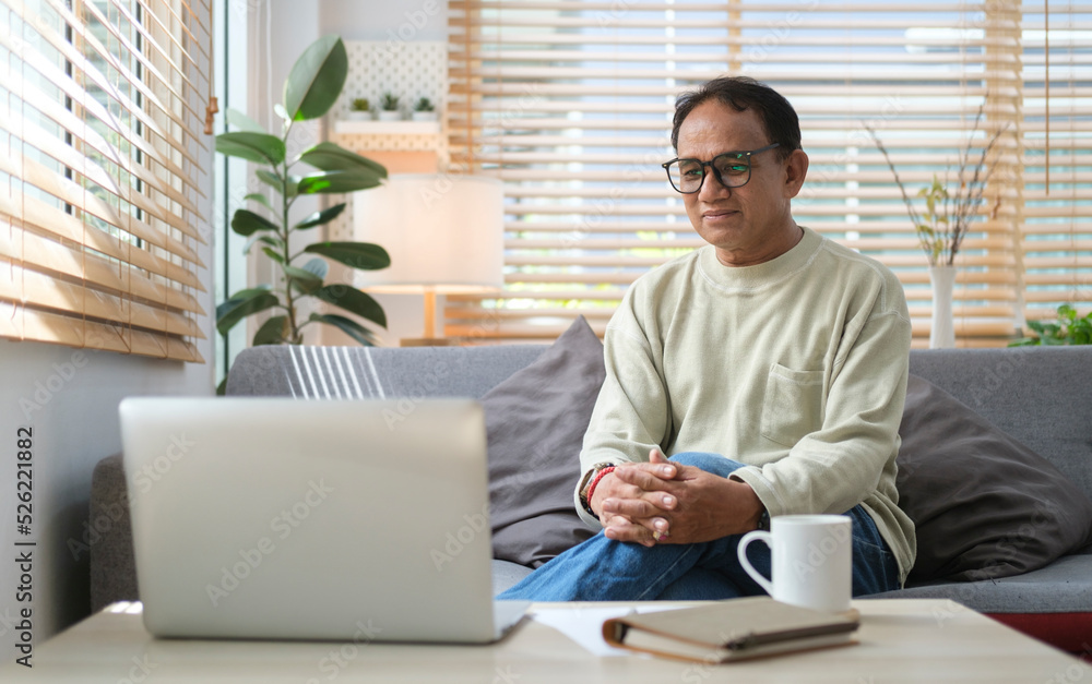 Retired man resting on couch in bright living room and reading online news on laptop at the morning.