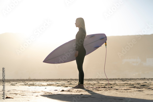 Surfer woman at the beach standing with her surfboard at sunset. Female surfer