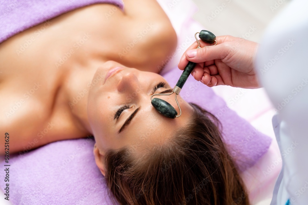 Fototapeta premium Facial massage. Side view european woman getting massage with jade face roller gouache in spa salon