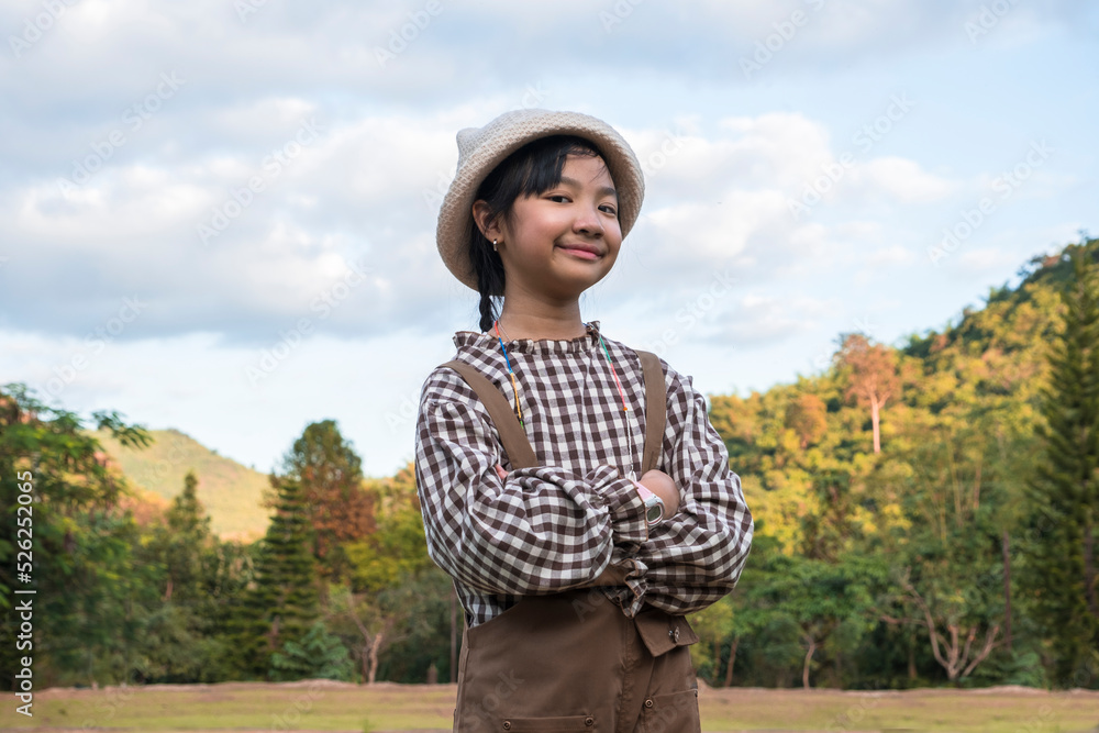 Asian girl in a brown overalls wearing a plaid shirt and hat is smiling in a farm with green trees.