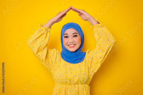 Cheerful young Asian woman in casual dress making rooftop home gesture with hands above head isolated on yellow background © Sewupari Studio