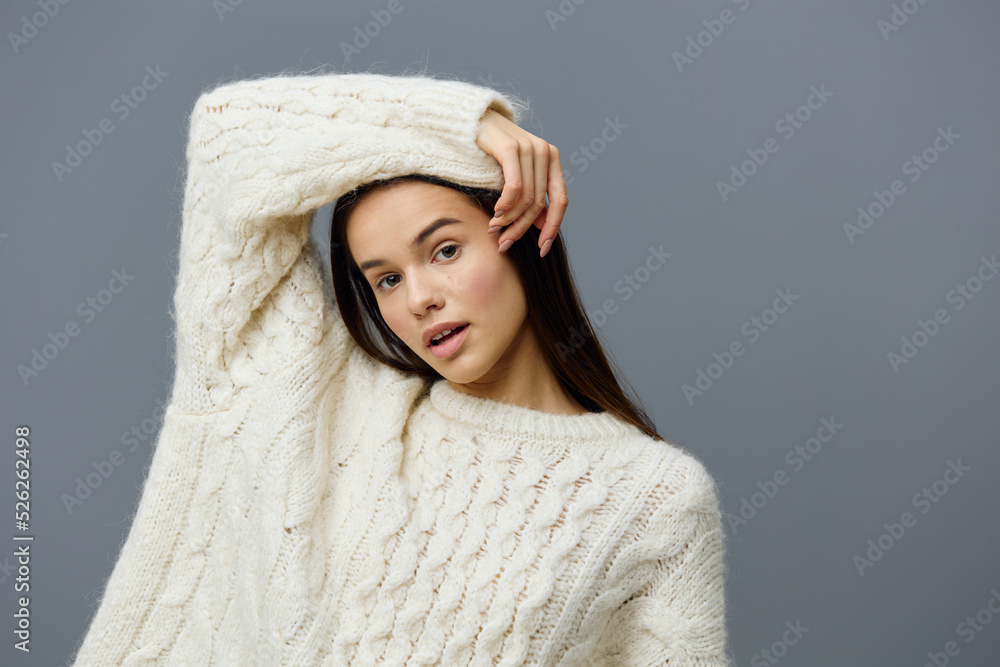 a beautiful, gentle, sophisticated woman with long hair stands in a stylish sweater on a gray studio background touching her head with her hand