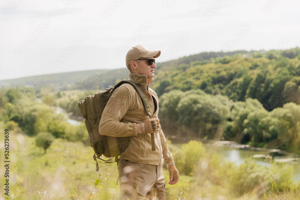 Masculine leisure concept: strong male figure with backpack stands on beautiful natural summer landscape outdoors near river
