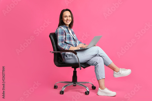 Mature woman with laptop sitting in comfortable office chair on pink background
