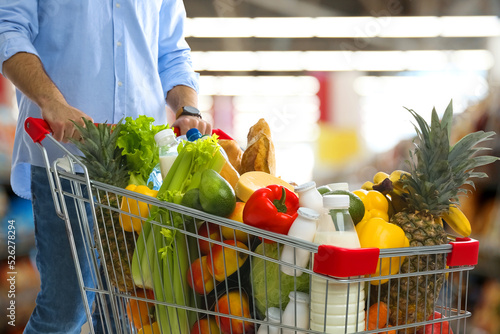 Man with shopping cart full of groceries in supermarket, closeup photo