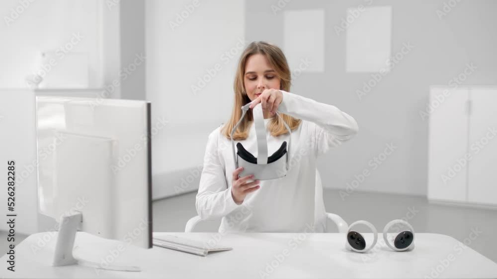 Young woman in white clothes put on vr headset sitting at desk in white room Young woman in white clothes put on vr headset sitting at desk in white room