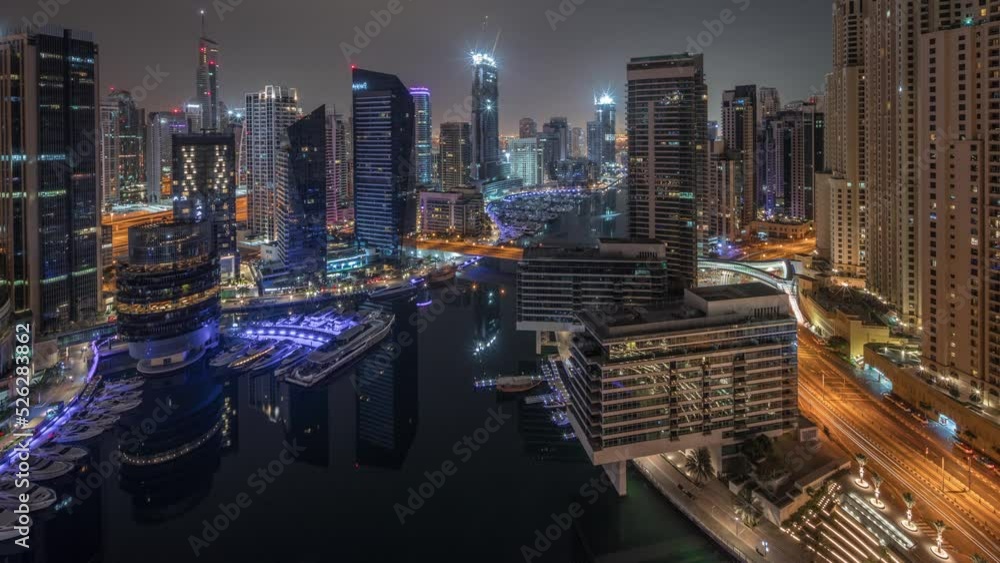 Aerial panoramic view to Dubai marina illuminated skyscrapers around canal with floating yachts timelapse during all night and lights turning off. White boats are parked in yacht club Aerial panoramic view to Dubai marina illuminated skyscrapers around canal with floating yachts timelapse during all night and lights turning off. White boats are parked in yacht club