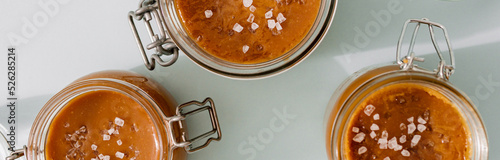 Salted caramel in glass jars, banner horizontal, top view. Brown caramel or condensed milk with sea salt crystalls, shoot in bright or hard light. Light neutral background photo