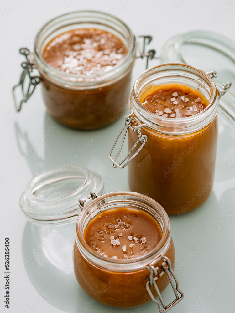 Salted caramel in glass jars. Brown caramel or condensed milk with sea salt crystalls, shoot in bright or hard light. Light neutral background. Vertical Salted caramel in glass jars. Brown caramel or condensed milk with sea salt crystalls, shoot in bright or hard light. Light neutral background. Vertical