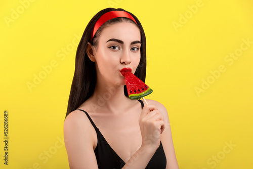 Photo of a beautiful woman with a watermelon on a stick posing on a yellow background.