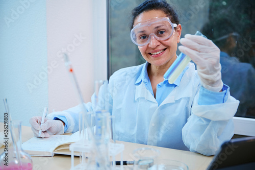 Positive multi-ethnic woman  scientist  medical researcher in safety labwear  holds test tubes with chemical substances  makes notes on nopetapd and smiles looking at camera  in a science laboratory