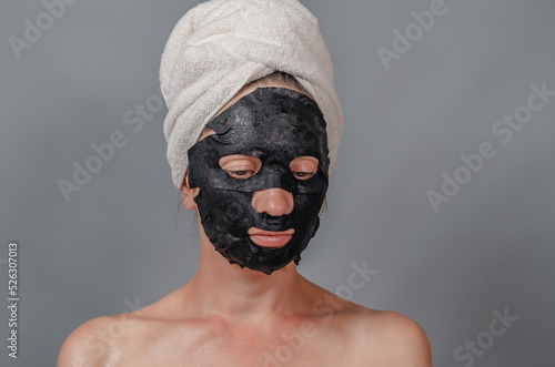 Portrait of a woman in a white towel on her head and a black fabric mask on her face. Moisturizing the skin. Gray background. © romankrykh