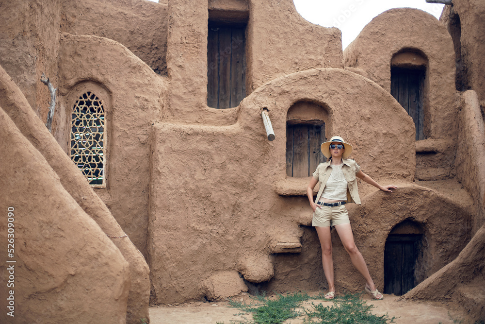 portrait of a traveler woman in a hat and sunglasses stands in an abandoned eastern city made of clay in summer. short sand shorts and jacket.