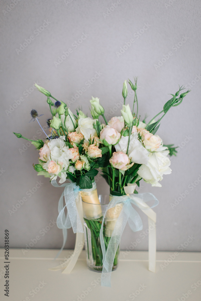 Two bouquets in light colors of white eustoma, white and cream roses, wrapped in blue ribbons, stand in a glass vase. Two bouquets in light colors of white eustoma, white and cream roses, wrapped in blue ribbons, stand in a glass vase.