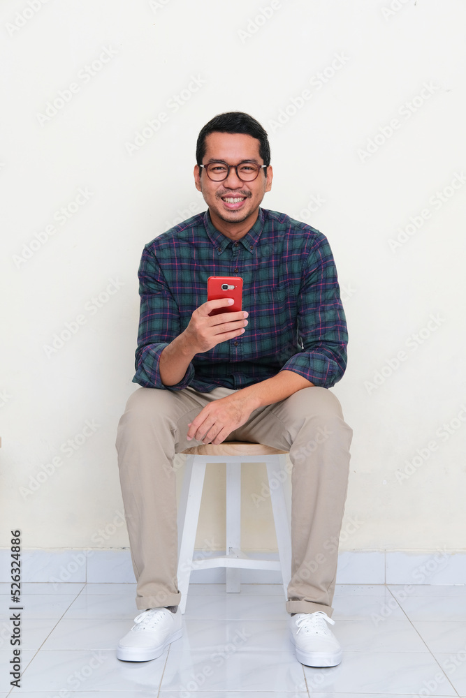 Asian man sitting in a bench smiling to camera while holding handphone Asian man sitting in a bench smiling to camera while holding handphone