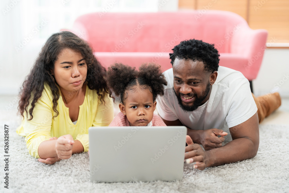 African American family laying down on the floor and looking at laptop with daughter with smile in the living room. Happy family day on vacation. Kid feeling boring with online learning African American family laying down on the floor and looking at laptop with daughter with smile in the living room. Happy family day on vacation. Kid feeling boring with online learning