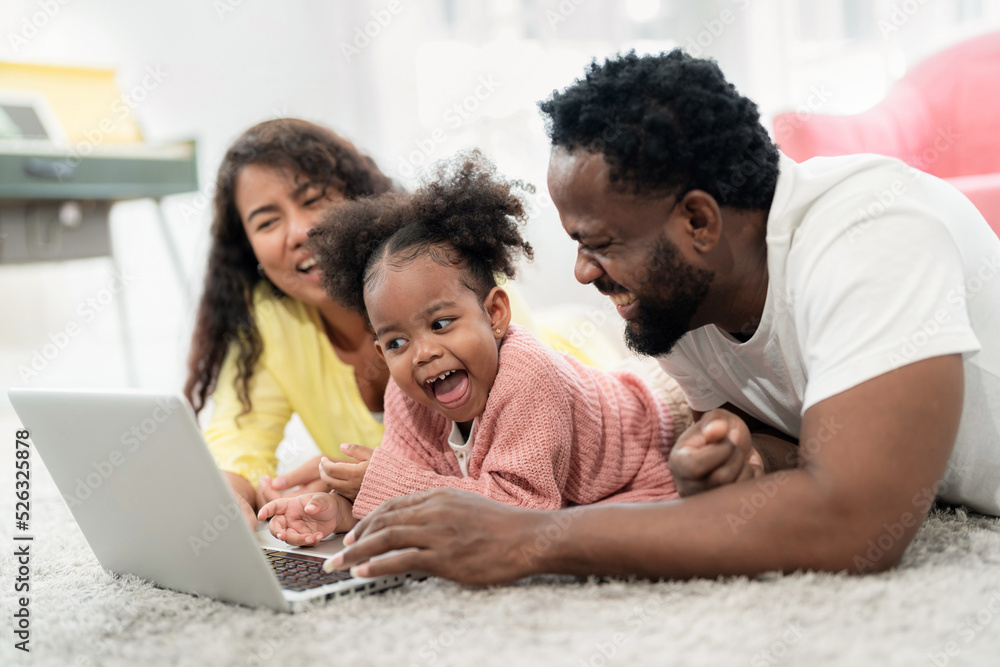 African American family laying down on the floor and looking at laptop with daughter with smile in the living room. Happy family day on vacation. Kid feeling happy and fun during playing game. African American family laying down on the floor and looking at laptop with daughter with smile in the living room. Happy family day on vacation. Kid feeling happy and fun during playing game.