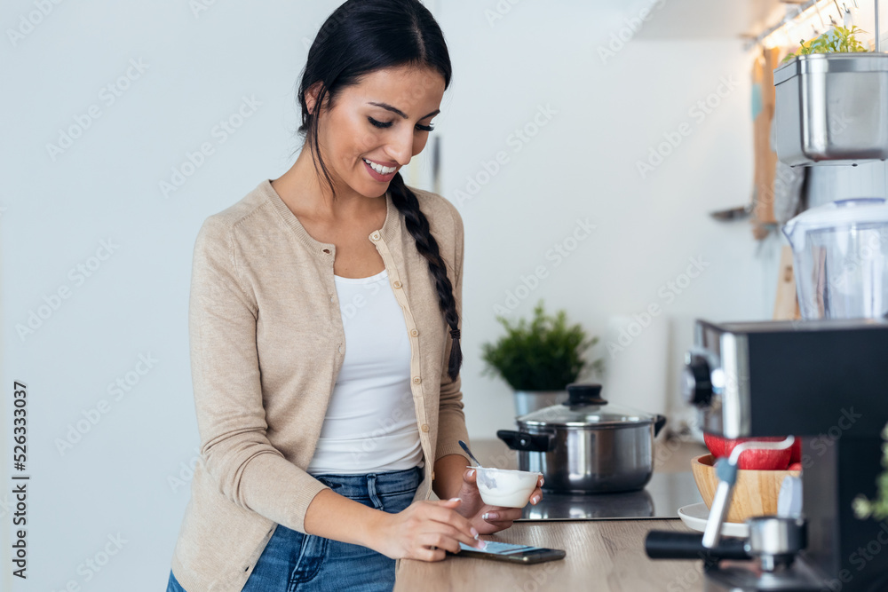 Pretty woman sending messages with mobile phone while eating yogurt int he kitchen at home. Pretty woman sending messages with mobile phone while eating yogurt int he kitchen at home.