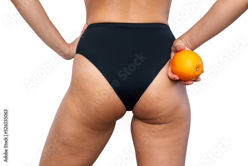 Young woman with cellulite and white stretch marks from a weight loss or weight gain on thighs stands with her back holding orange in her hand isolated on a white background. Excess weight, overweight