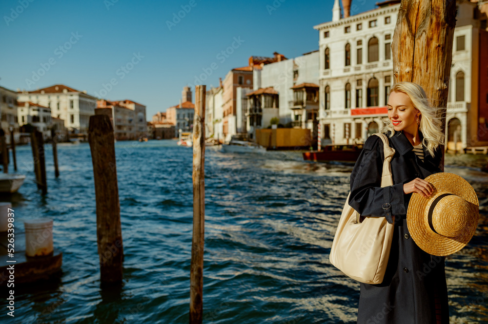 Elegant happy smiling traveler woman wearing blue trench coat, holding straw hat, with big hobo bag,  posing in street of Venice. Fashion, travel, lifestyle conception. Copy, empty space for text
