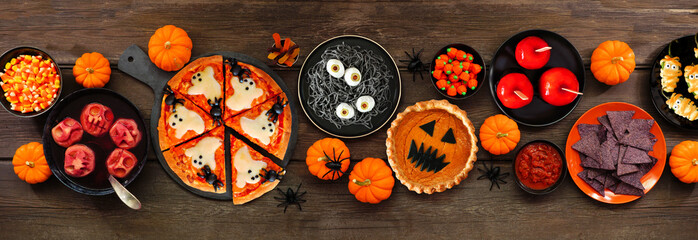 Fun Halloween dinner party table scene over a dark wood banner background. Above view. Pizza, jack o lantern pumpkin pie, candy apples, eyeball spaghetti, snacks and spooky punch.