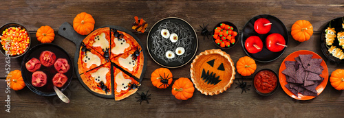 Fun Halloween dinner party table scene over a dark wood banner background. Above view. Pizza, jack o lantern pumpkin pie, candy apples, eyeball spaghetti, snacks and spooky punch. #526343490