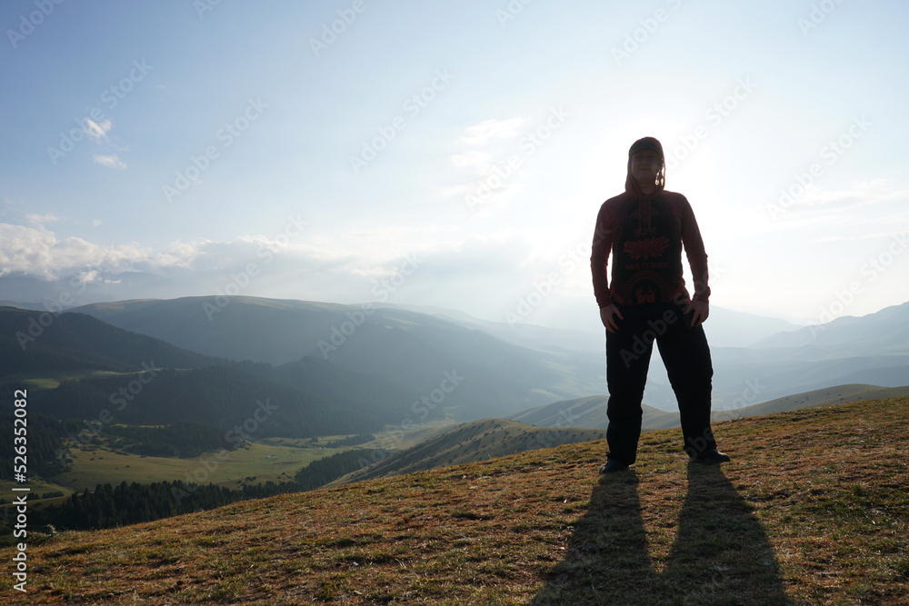 A guy on the edge of the hill admires the view. In the distance you can see high mountains and green hills covered with forest. Wide margins. Clouds are visible and sun is shining brightly