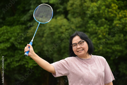 Portrain of asian middle aged woman holding badminton racket, standing and smiling in the green field to play outdoor badminton, concept for to be strong and healthy woman by exercising afterwork. © Sophon_Nawit