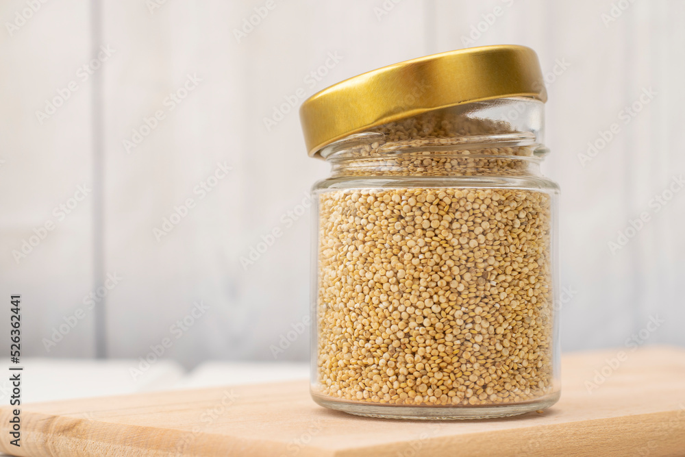Glass jar with quinoa seeds on a wooden board on a light background close-up, soft focus, space for text. Uncooked white quinoa seeds in a glass jar close-up. Glass jar with quinoa seeds on a wooden board on a light background close-up, soft focus, space for text. Uncooked white quinoa seeds in a glass jar close-up.