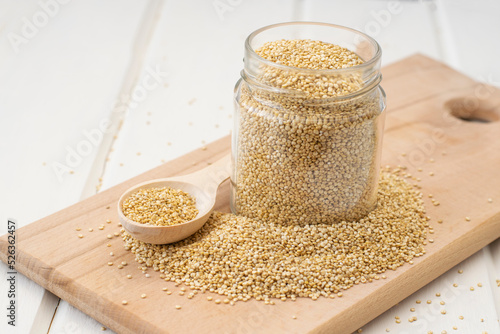 Glass jar with quinoa seeds and white uncooked quinoa seeds scattered around the jar on a wooden board on a light background close-up, soft focus.