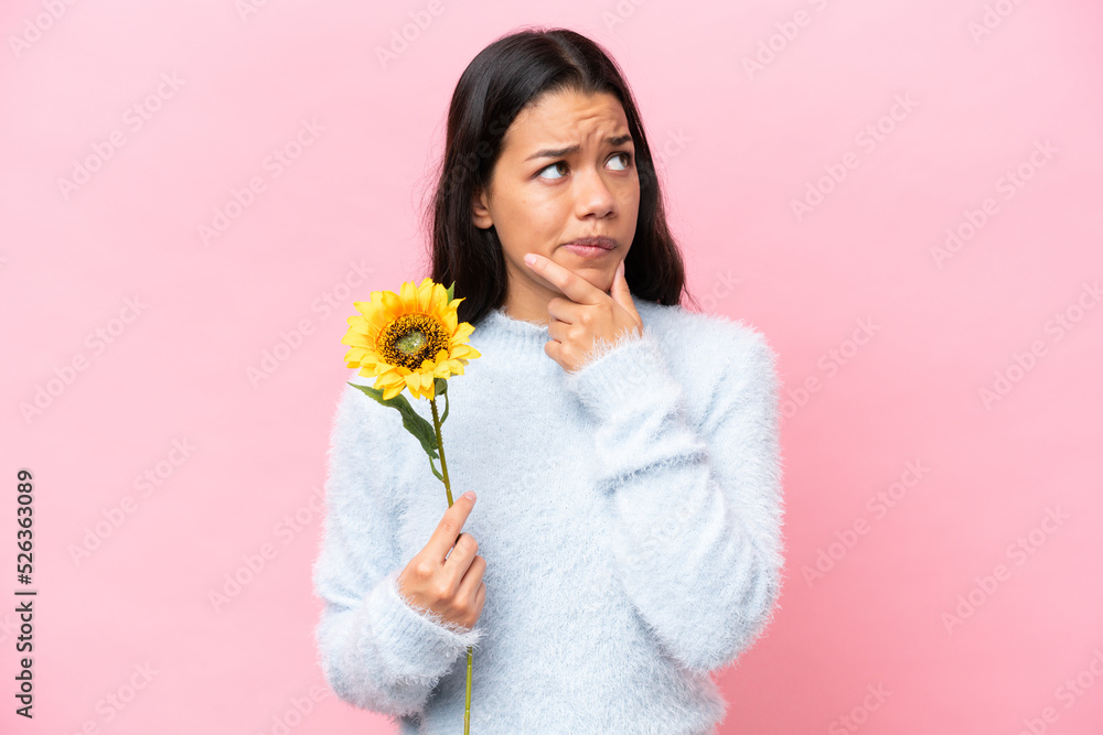 Young Colombian woman holding sunflower isolated on pink background having doubts and thinking Young Colombian woman holding sunflower isolated on pink background having doubts and thinking